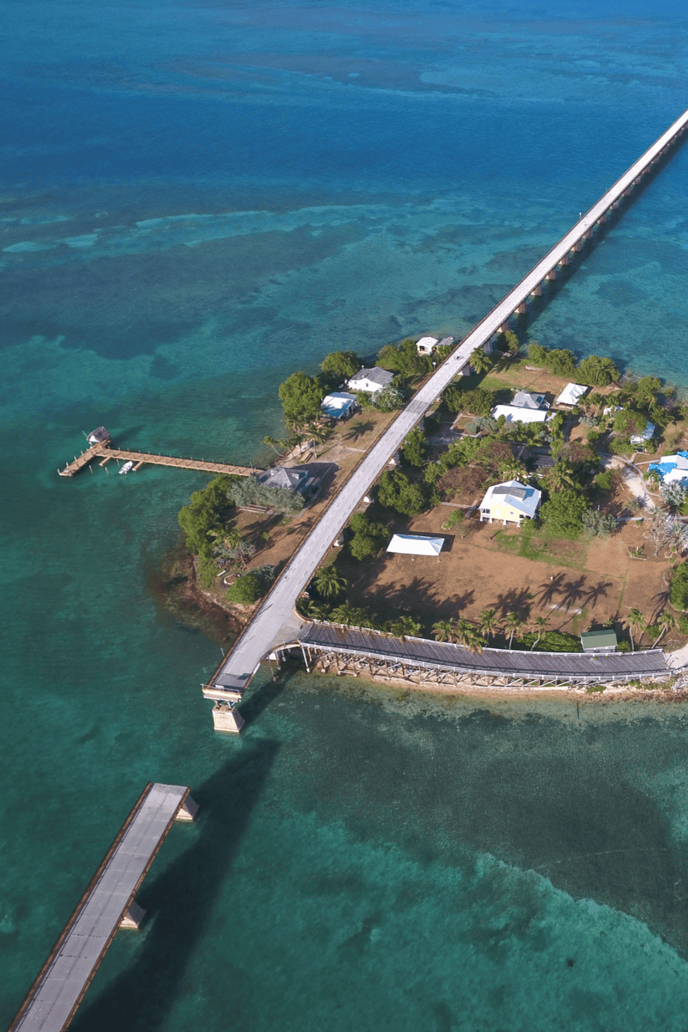 Aerial view of a coastal bridge leading to a small island with houses and a pier, surrounded by turquoise waters and lush greenery.