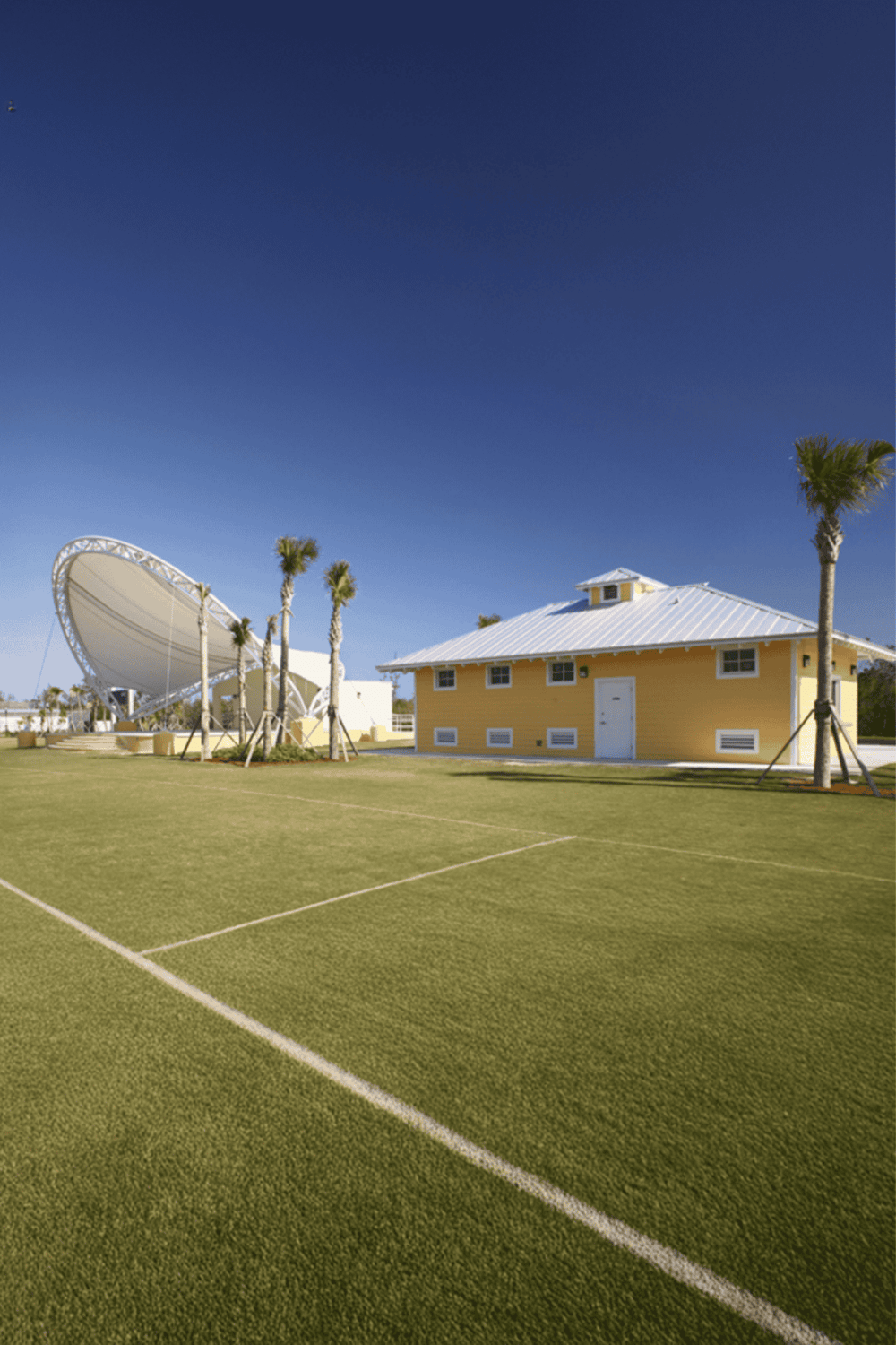 Shaded sports field near a yellow building with satellite dish, palm trees, clear sky, sunny weather, and outdoor recreation area.