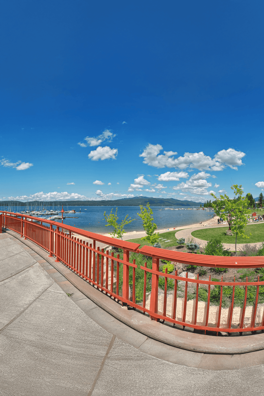 Vibrant waterfront park with boat marina, walking trail, and scenic mountain views under a bright blue sky.