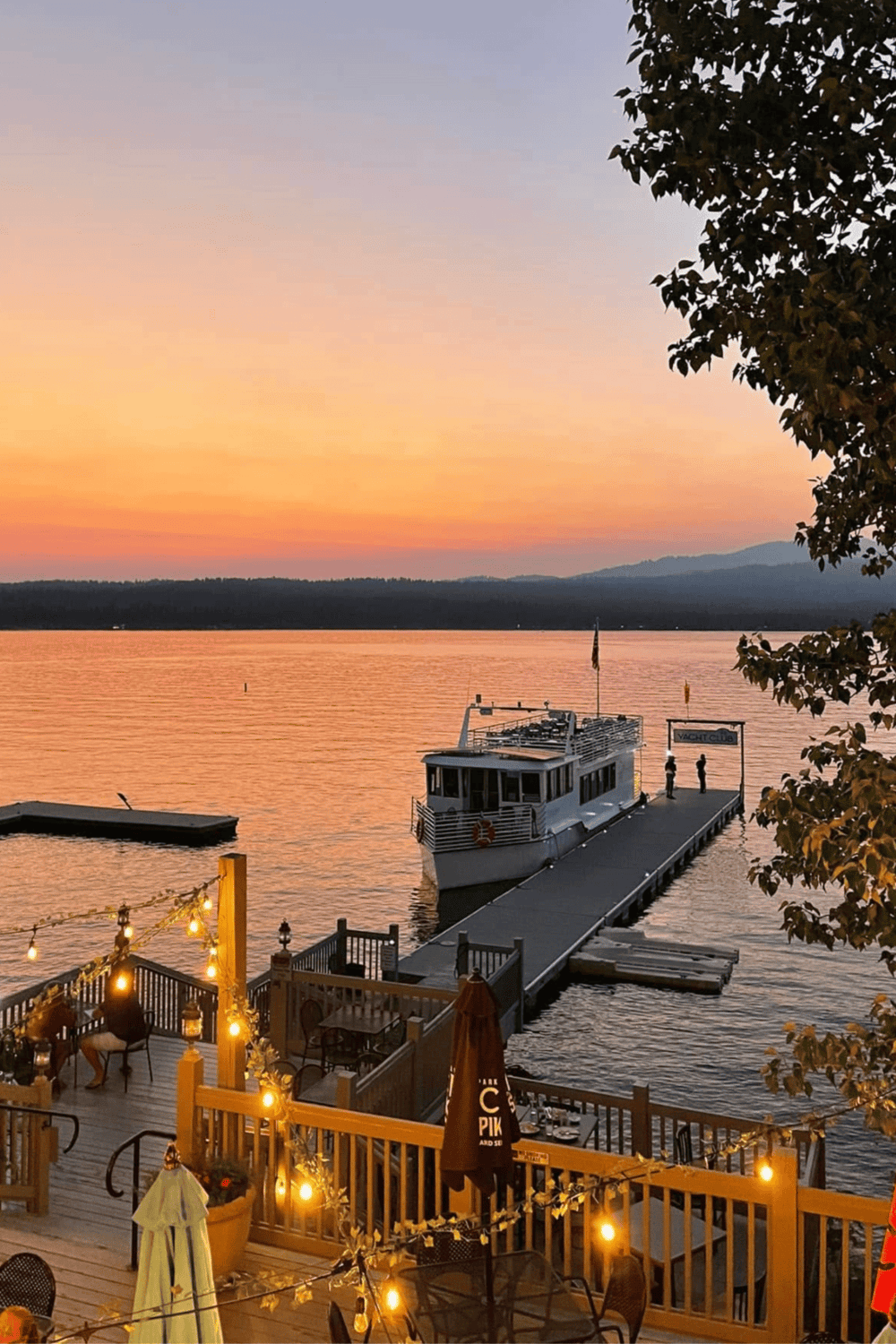Serene lake dock at sunset with boat, outdoor seating, and warm string lights, perfect for lakeside dining and sunset views.