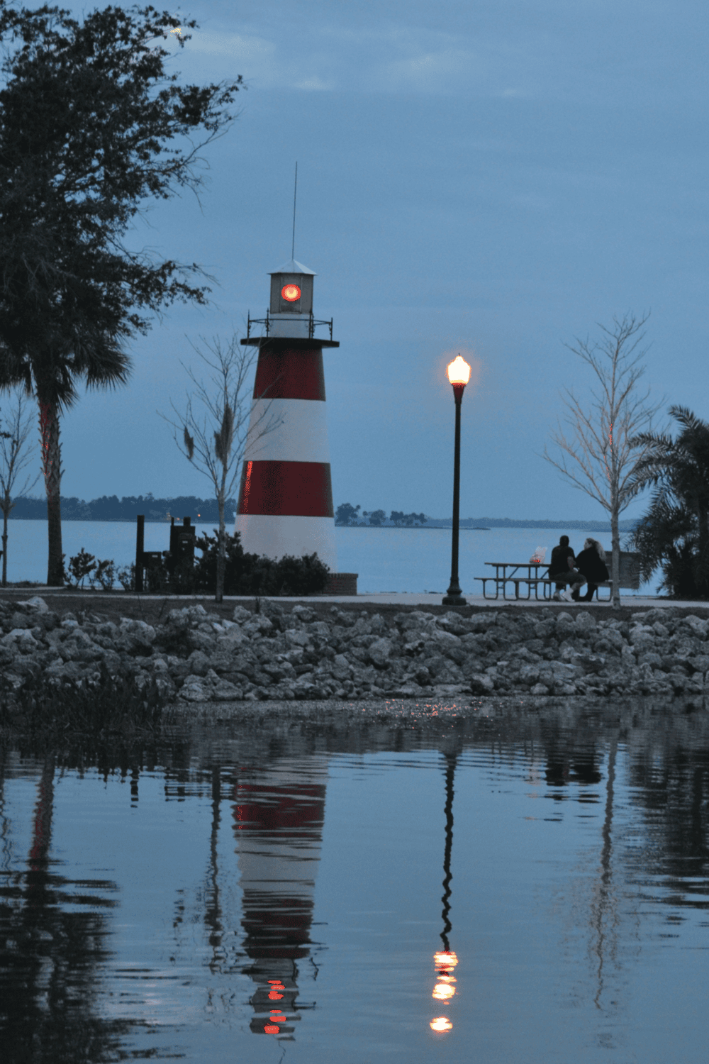Lighthouse by the waterfront during evening, with two people sitting at a picnic table nearby.