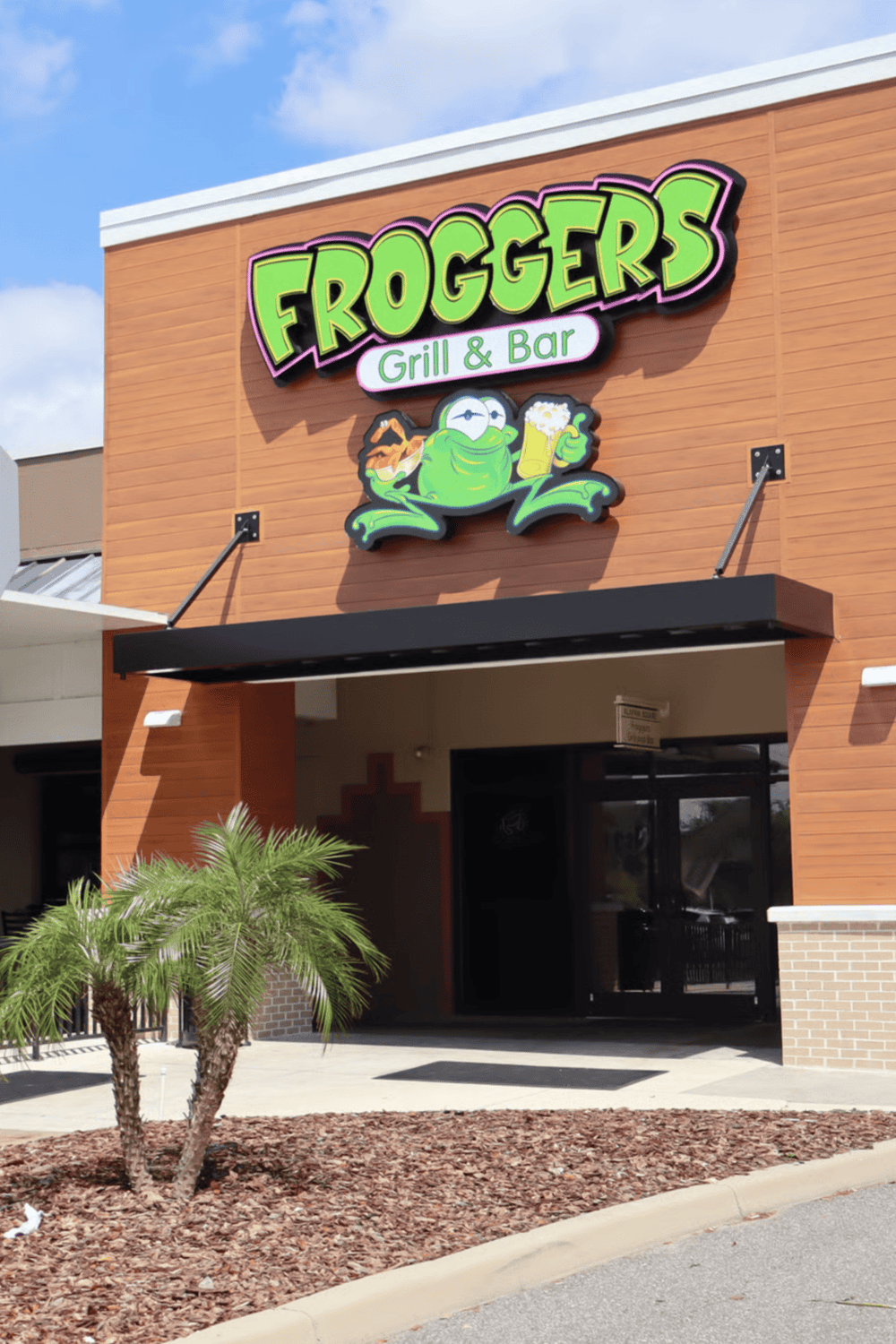 Brightly lit Froggers Grill & Bar restaurant sign with cartoon frog mascot, colorful logo, inviting exterior.