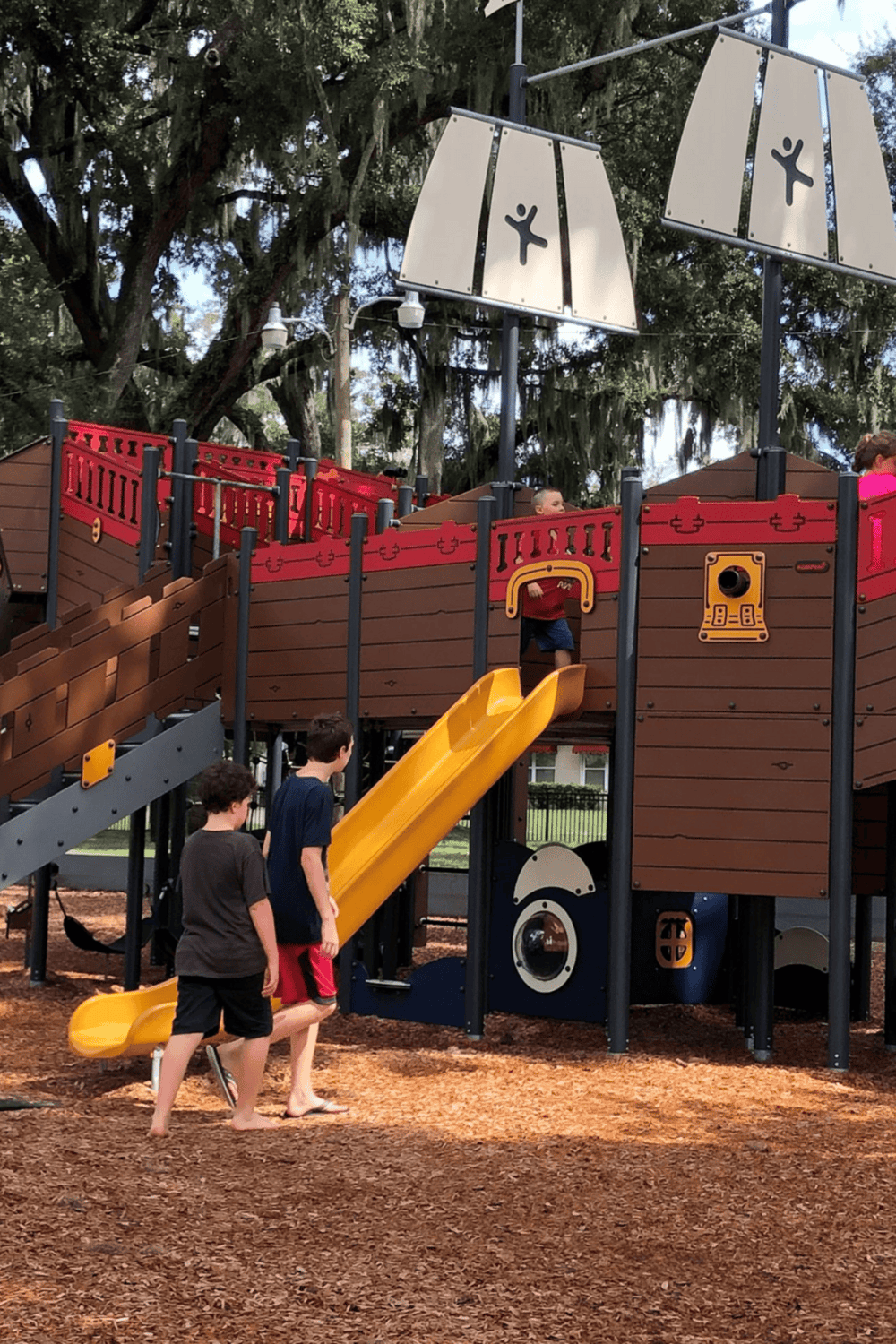 Kids playing on a pirate ship playground with a yellow slide, surrounded by trees and mulch, at QuestForDirections park.