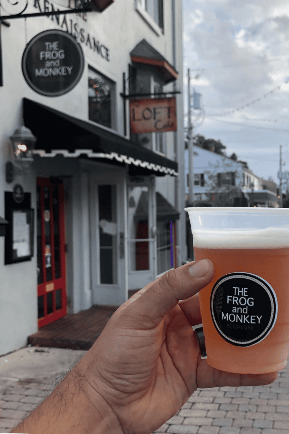 Cozy pub exterior with a hand holding a craft beer at The Frog and Monkey restaurant and pub. Ideal for casual dining and drinks.