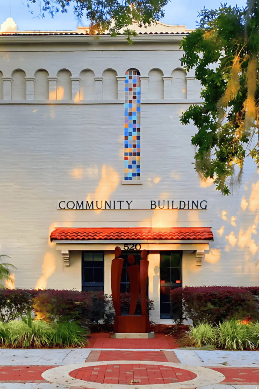 - White community building with colorful mosaic art and sculpture in front.