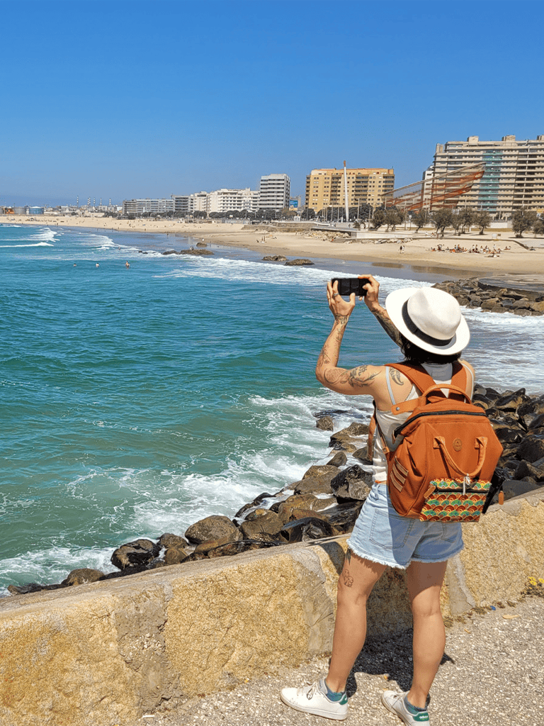 Vibrant woman taking photo at sunny beach with city skyline in background.