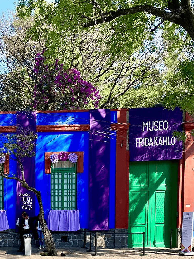Vibrant colorful exterior of Museo Frida Kahlo with purple, green, and blue accents, surrounded by lush trees and flowers.