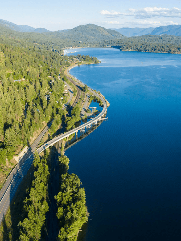 Lush green forest and highway along a large blue lake with mountain backdrop.