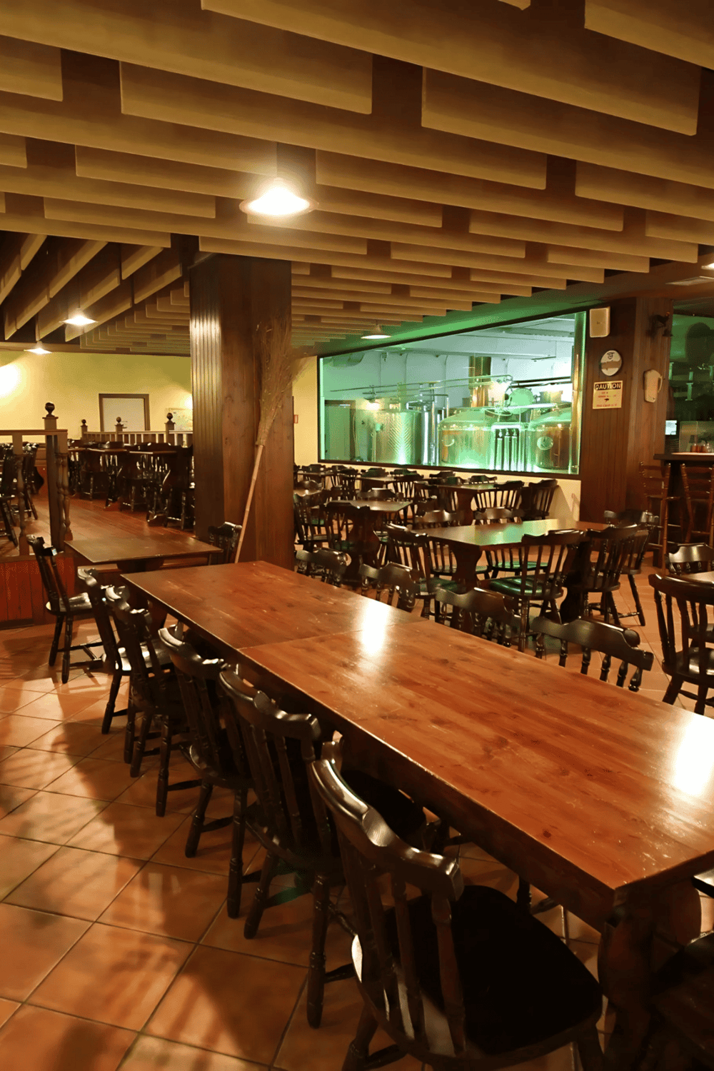 Cozy restaurant interior with wooden tables and chairs, brewery tanks visible through glass wall.
