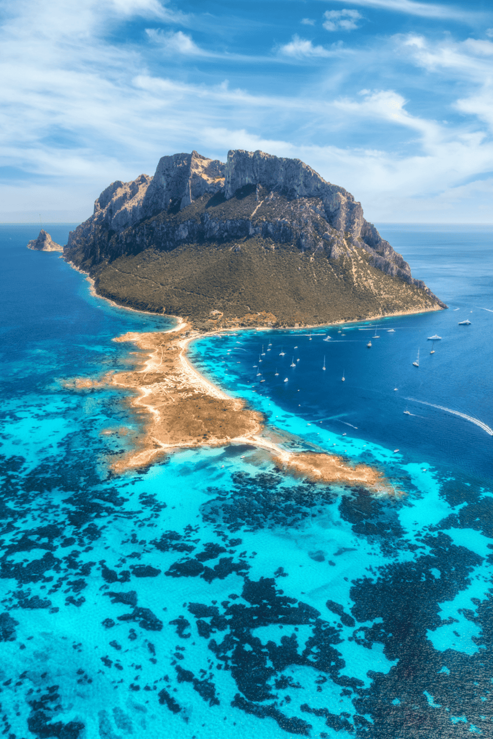 Aerial view of a scenic island with turquoise waters, mountain landscape, and boats anchored around the coast.