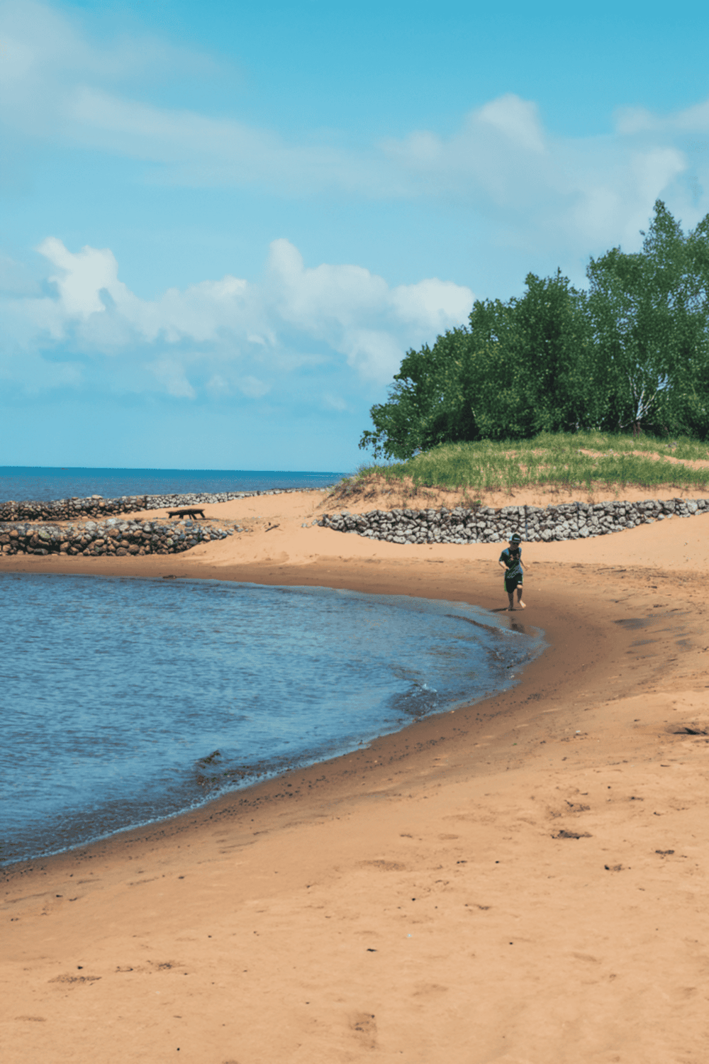 Seaside beach with rocks, trees, and person walking along shoreline, scenic coastal outdoor location.