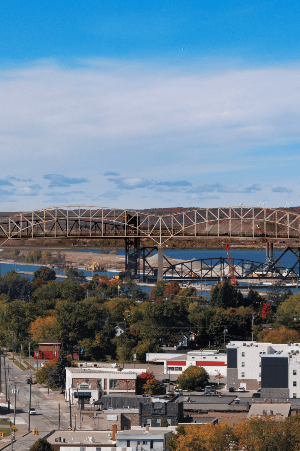 Suspension bridge over river in scenic cityscape with autumn foliage and rolling hills, showcasing urban development and natural beauty.