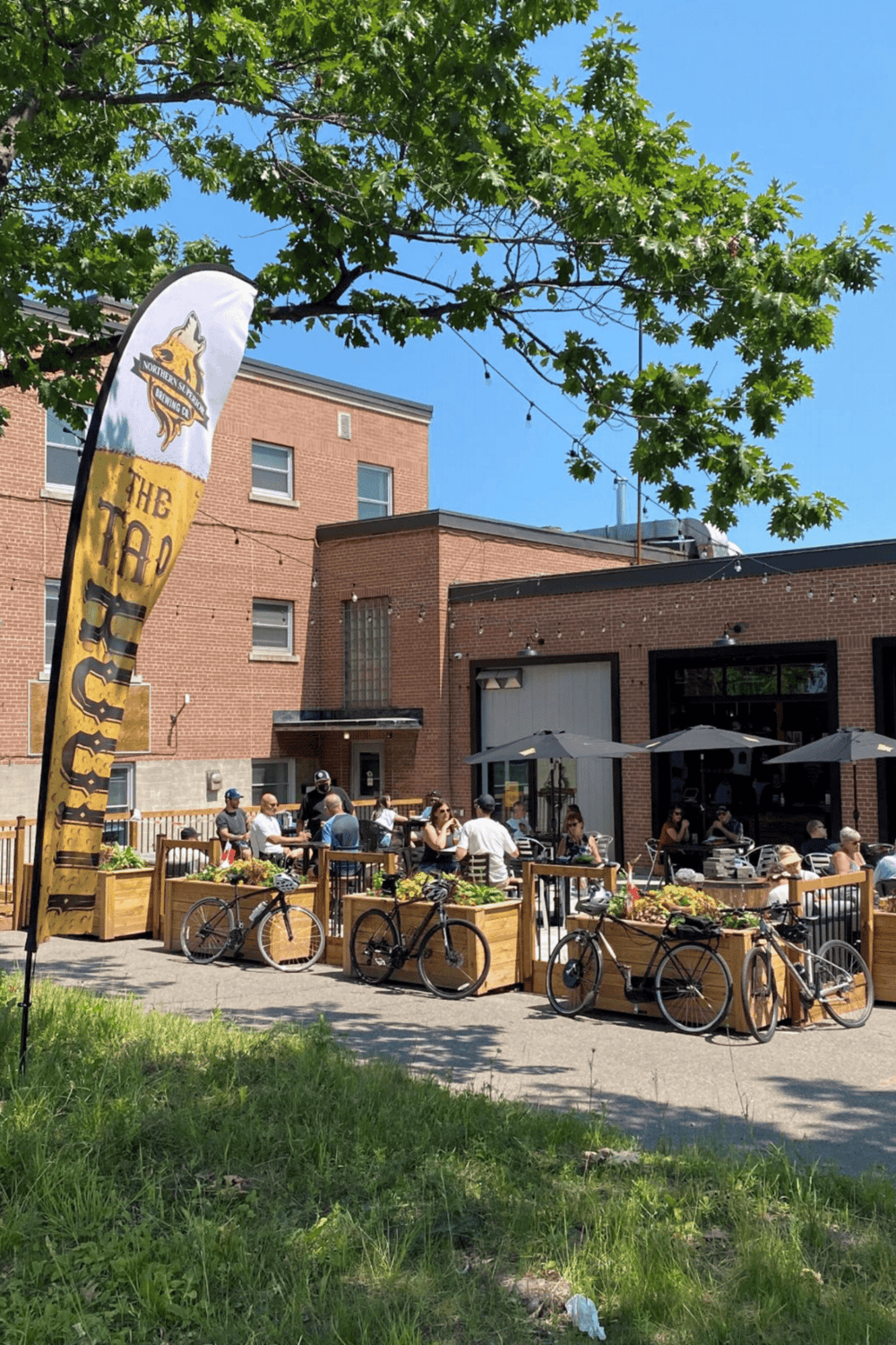 Bicycle outdoor dining area with people, brick building, trees, and sunny sky in the background.