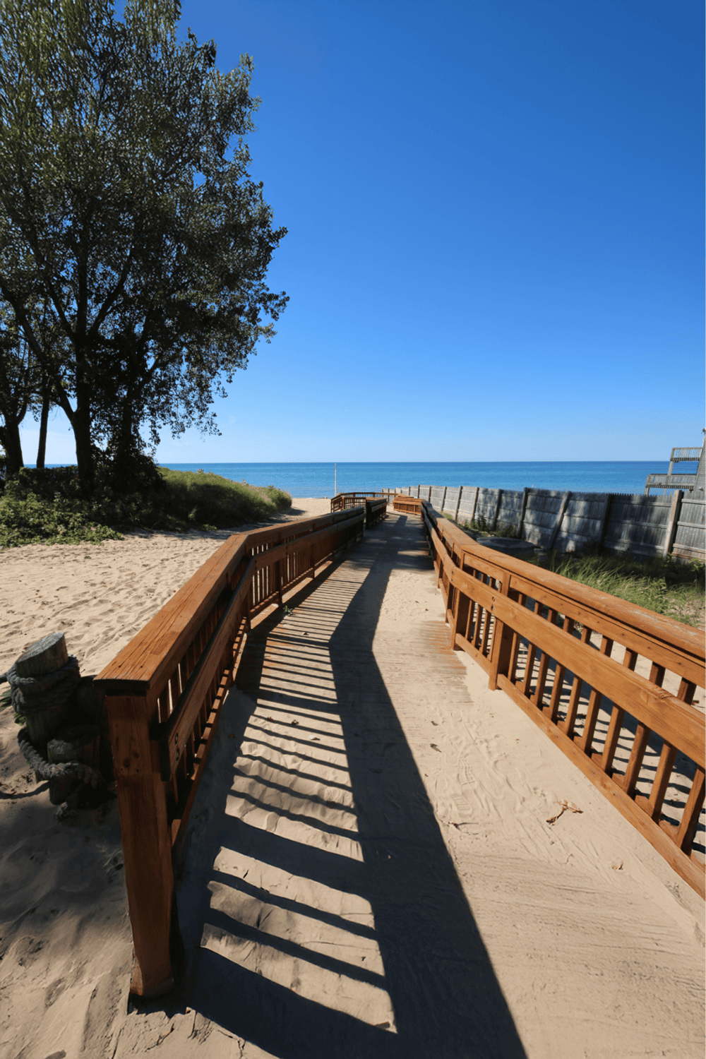 Beach walkway to ocean with wooden rails and sandy path, bright sunny day with clear blue sky.