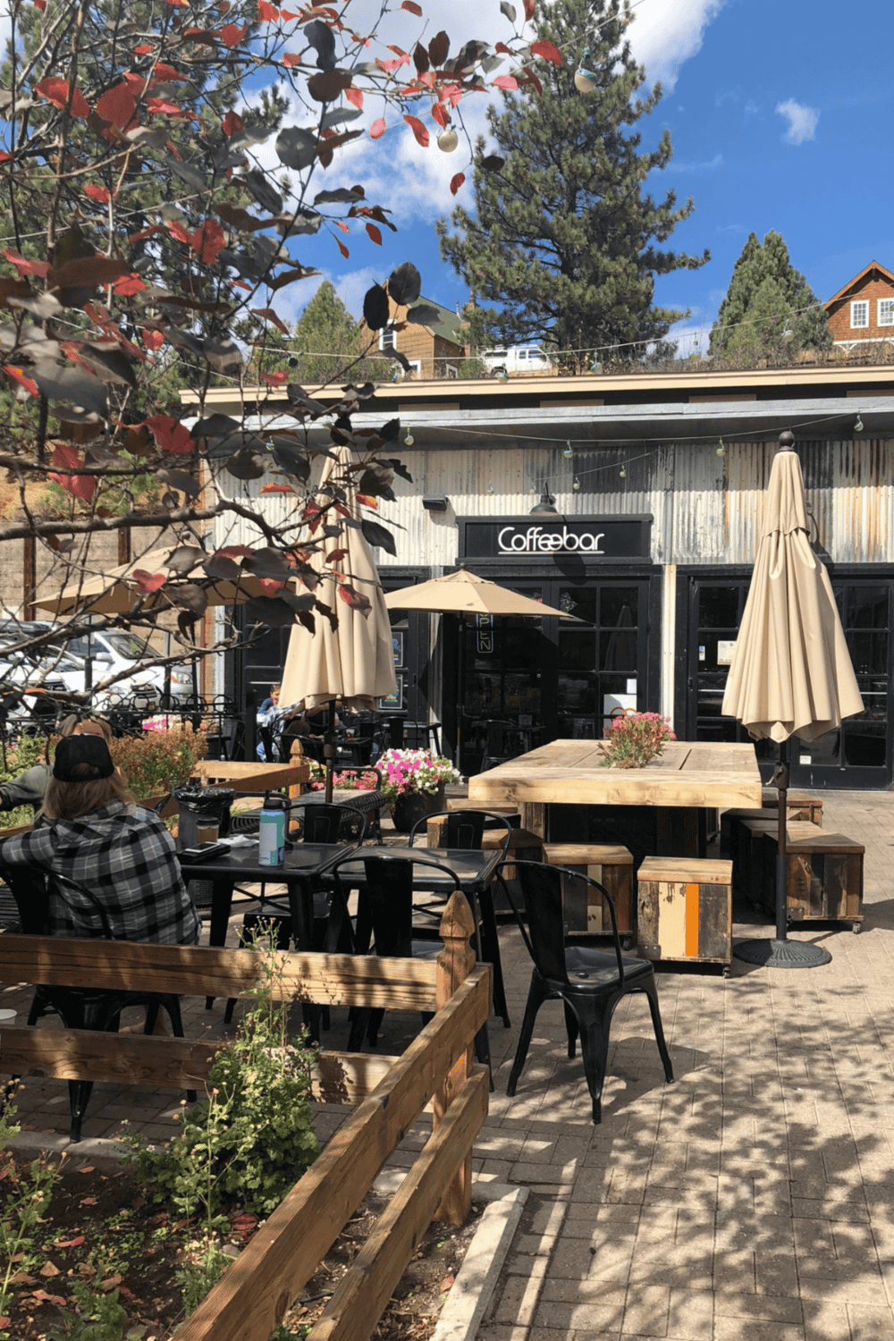 Outdoor coffee shop with seating under umbrellas, surrounded by trees and flowers, in a scenic mountain town setting.