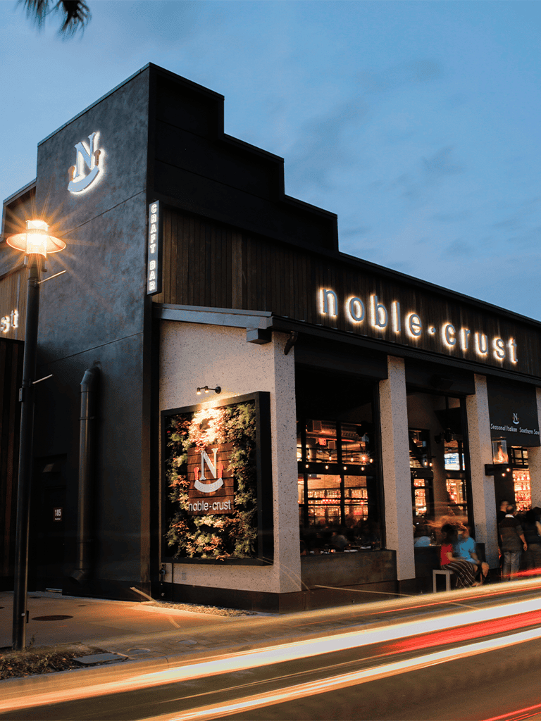 Modern Noble Crust restaurant exterior at dusk, illuminated signage, inviting dining atmosphere, urban setting.