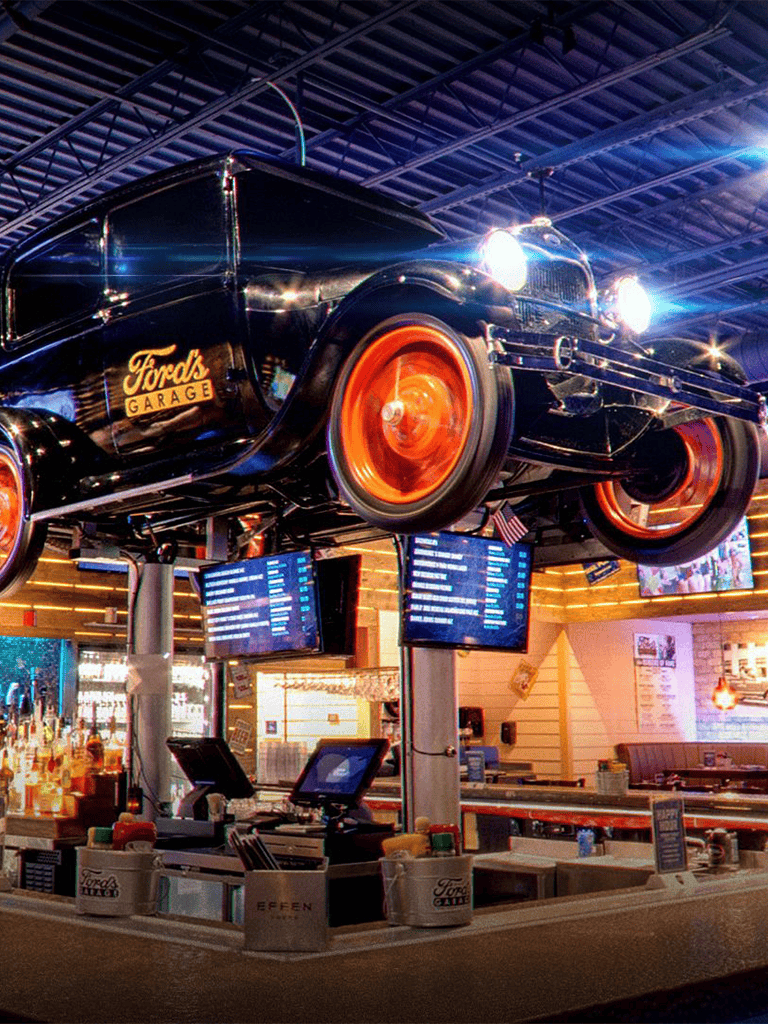 Ford car hanging at Ford's Garage, vintage ambiance, modern bar, flight information displays, vibrant lighting, automotive-themed decor.