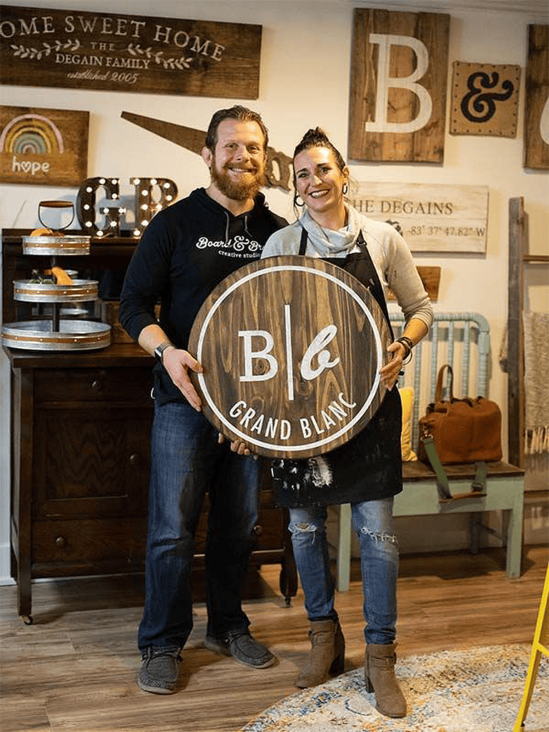 Cozy home decor with rustic wooden signs and cheerful entrepreneurs holding a "Board & Brush Grand Blanc" sign.