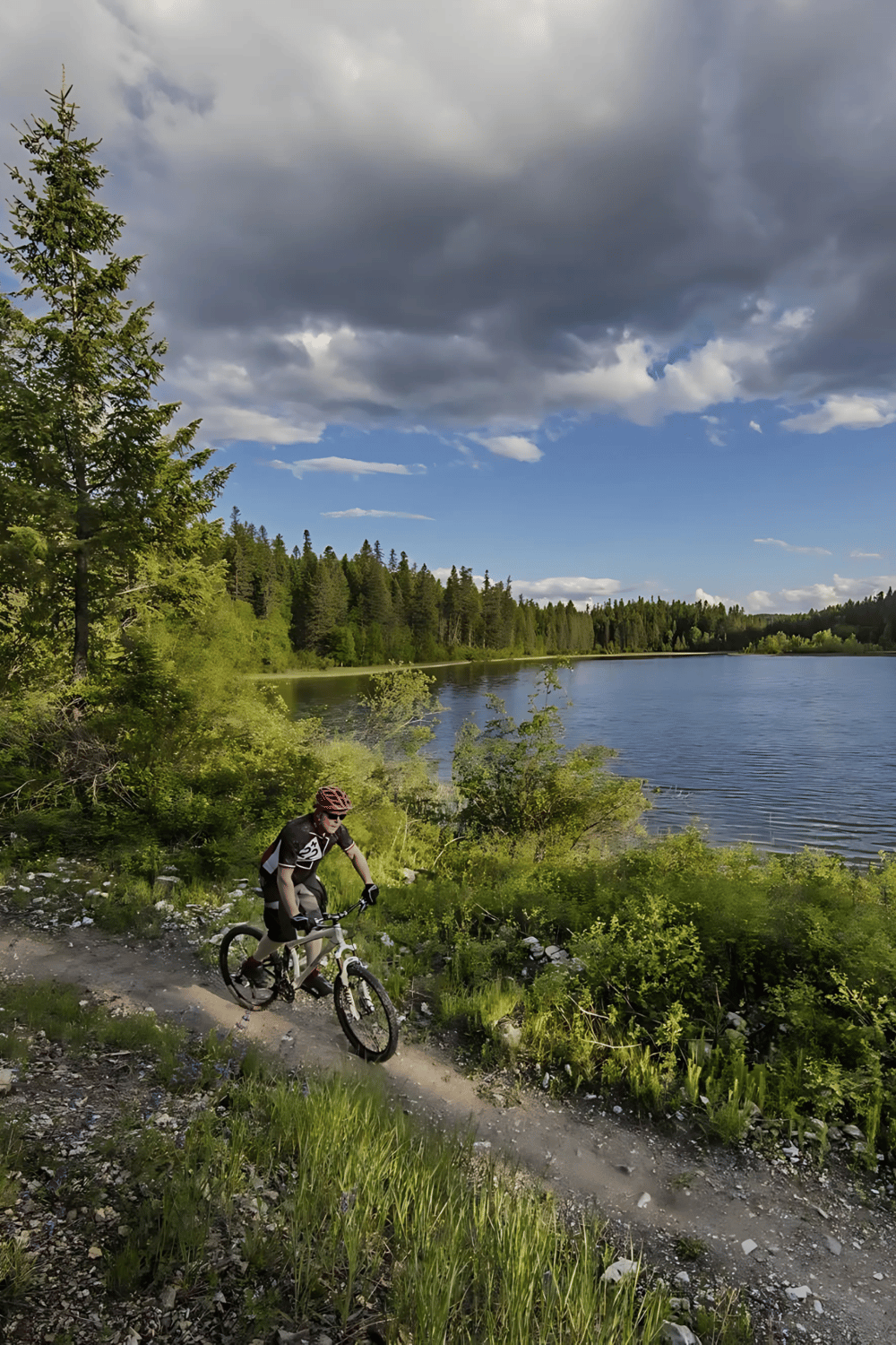 Scenic mountain biking along a forest trail beside a peaceful lake with dramatic cloud-filled sky.