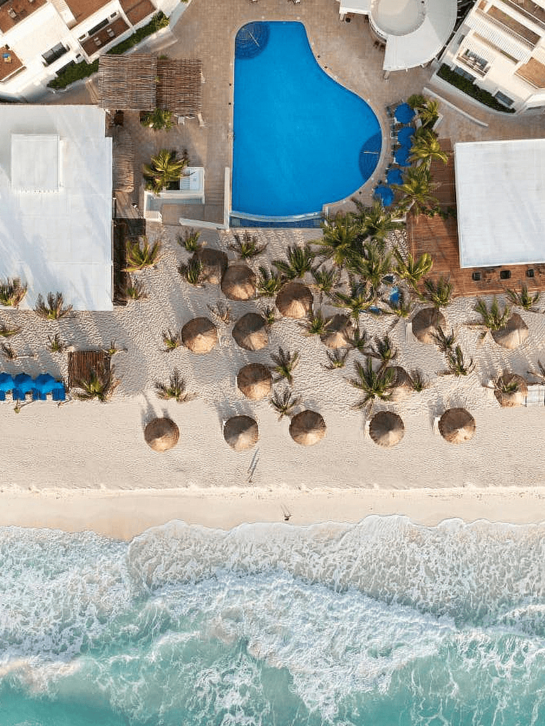 Beach resort with pool, palm trees, and straw umbrellas overlooking the ocean.
