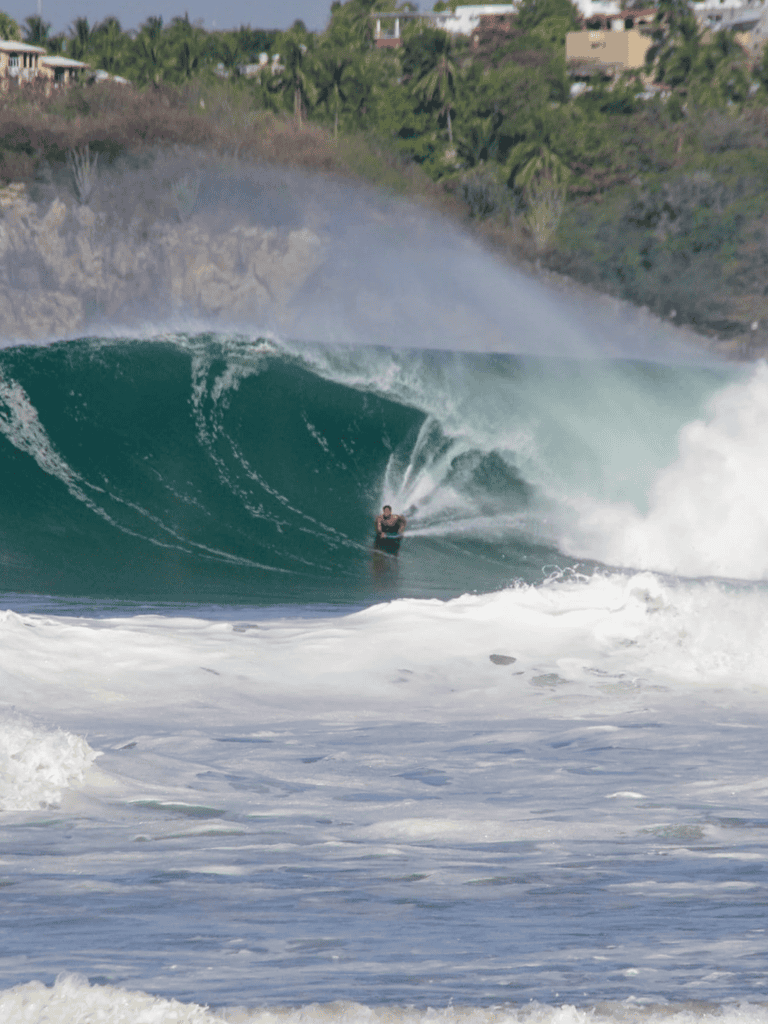 Vibrant surfer riding a large wave at a tropical beach with hillside homes in the background.