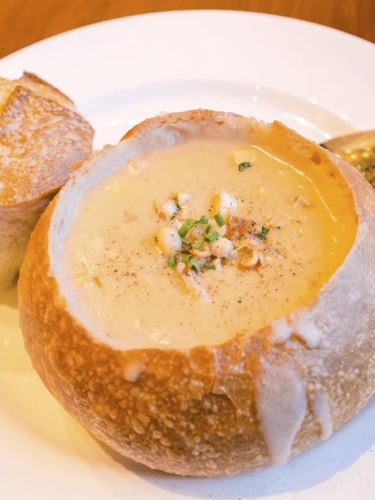 Creamy stuffed bread bowl with soup, topped with herbs, served with bread on a white plate.