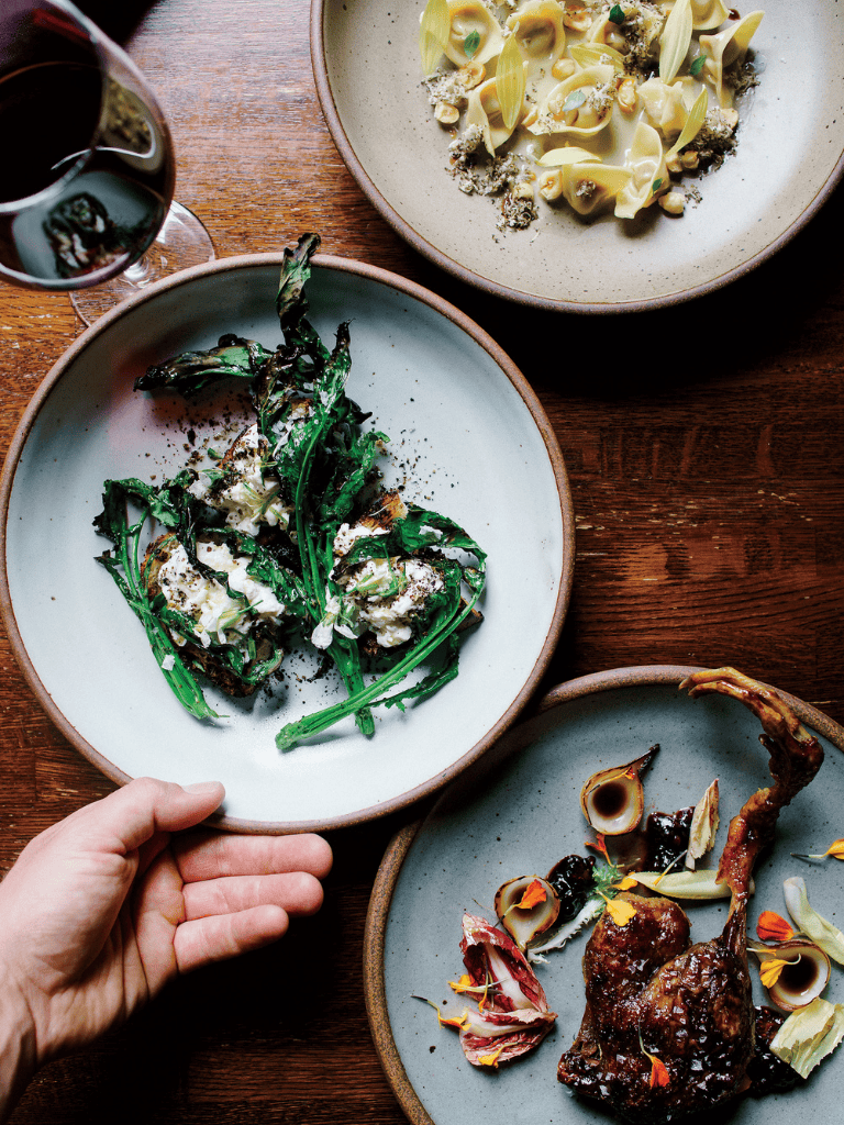 Creamy pasta with cheese, grilled greens, and roasted meat dishes on rustic wooden table.