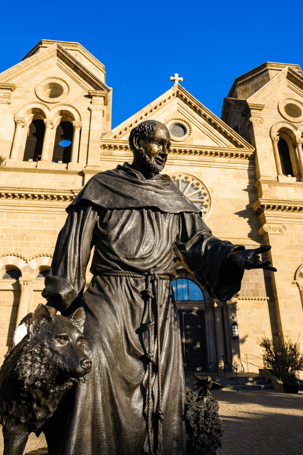 Bronze statue of Saint Francis of Assisi outside a historic church, symbolizing faith and compassion, with clear sky in the background.