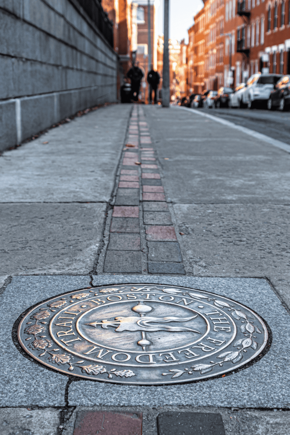 Historic Boston manhole cover on city sidewalk with brick street and pedestrians in the background.