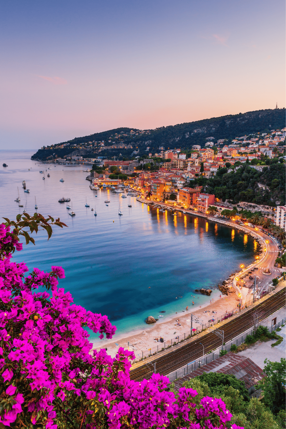 Stunning waterfront view of colorful seaside town with boats, hillside homes, and pink bougainvillea in the foreground.
