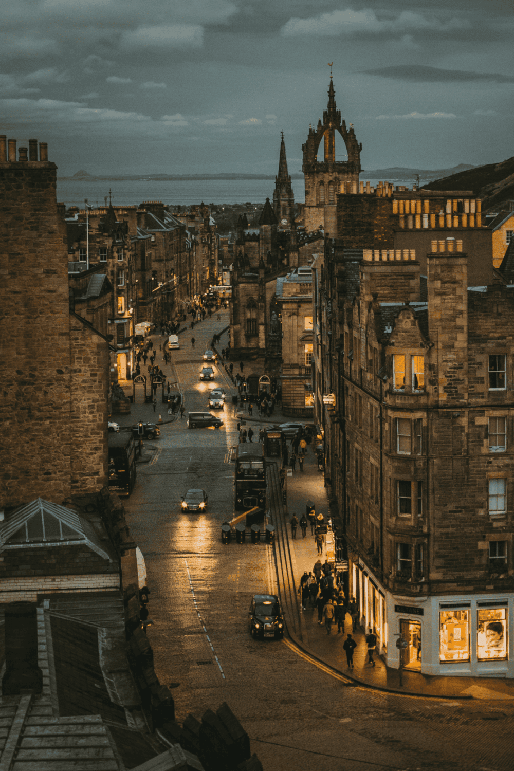 Victorian Edinburgh street at dusk with historic architecture, city lights, and scenic waterfront view.