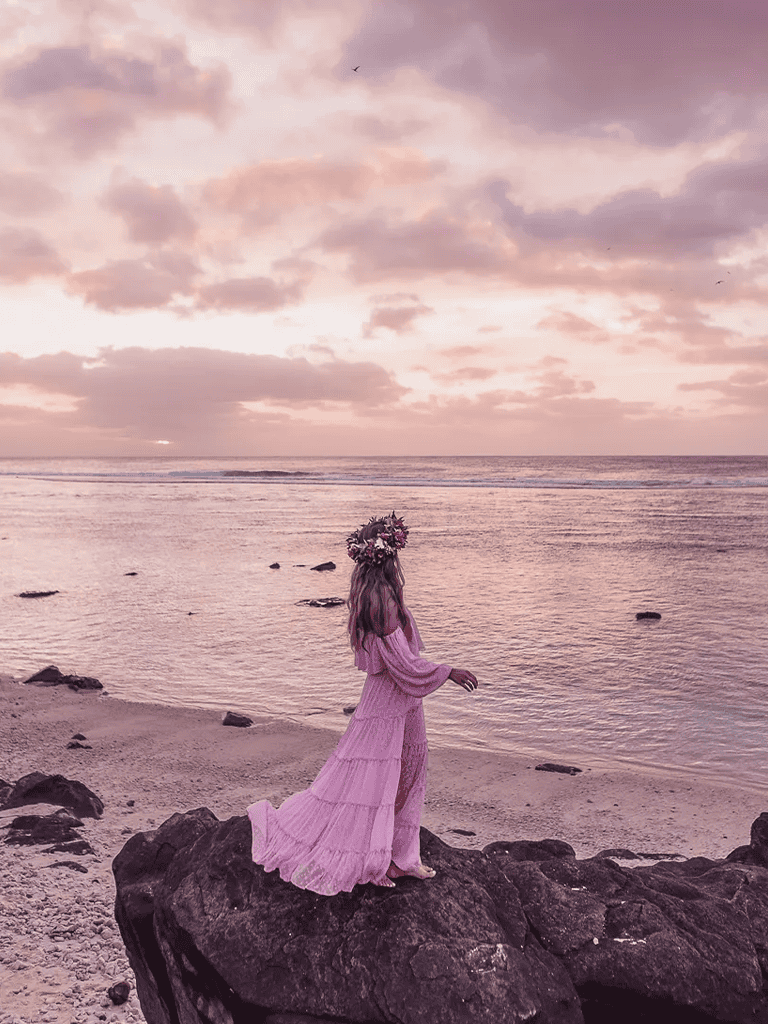 1. Woman in pink dress with floral crown on rocky beach at sunset, serene ocean view.