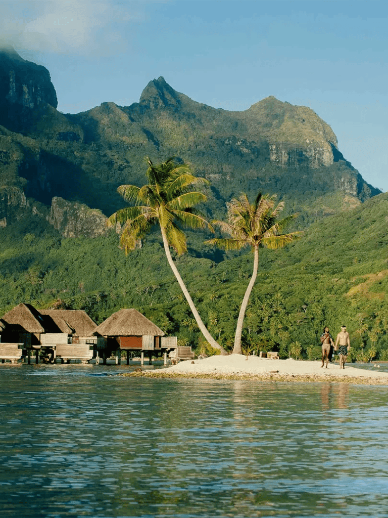Tropical island with palm trees, overwater bungalows, and lush green mountains in the background.