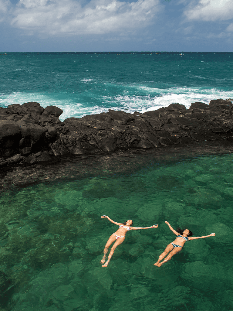 Relaxing ocean scene with women floating in clear green water near rocky shoreline, perfect for travel and leisure.