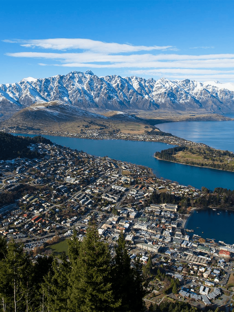 Snow-capped mountains over a city with lakes and lush greenery, scenic New Zealand landscape.