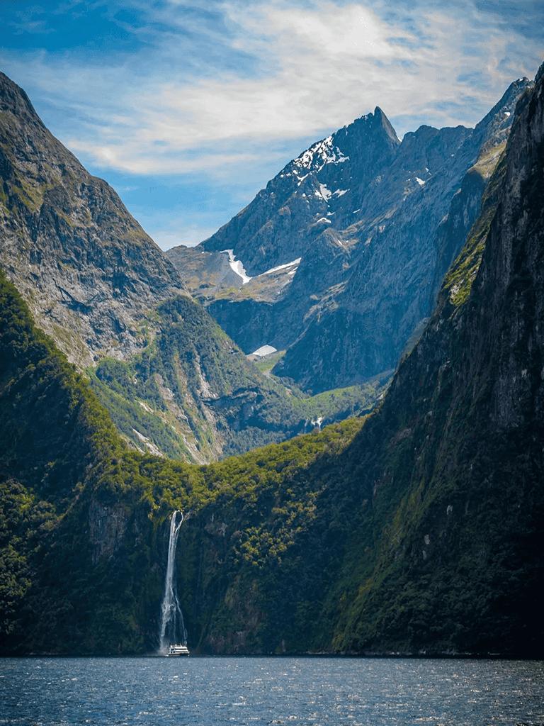 Breathtaking glacier-carved fjord with waterfall, towering mountains, lush greenery, and clear blue sky.