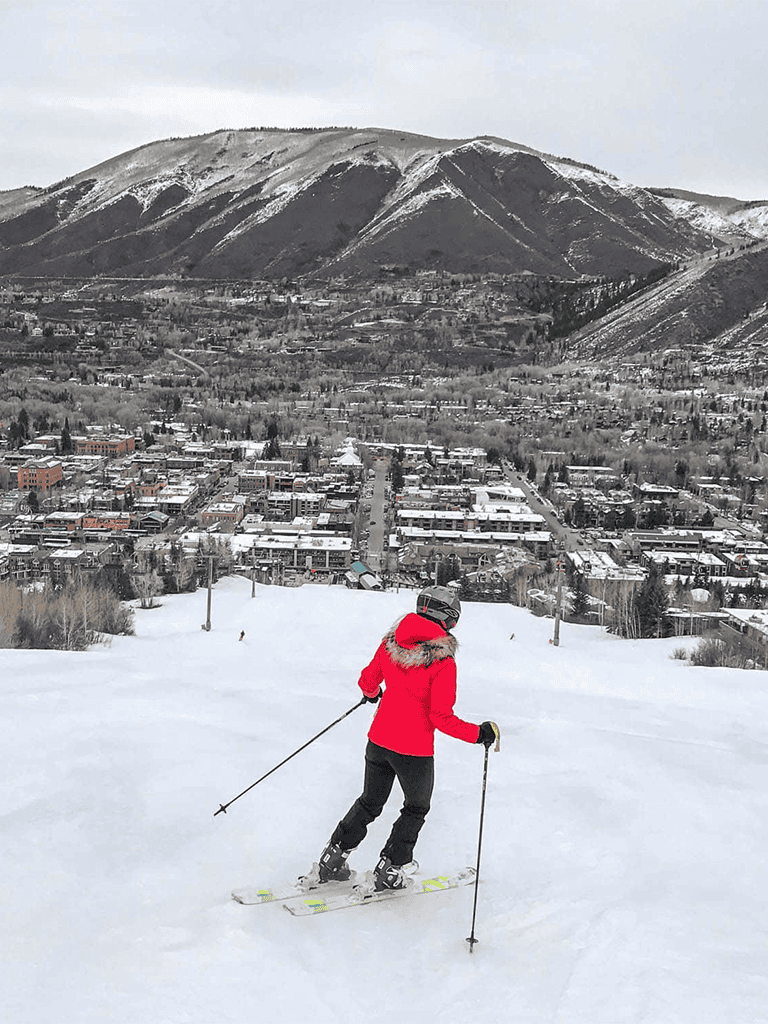 Snow skier in red jacket skiing down slope with mountain town in background.