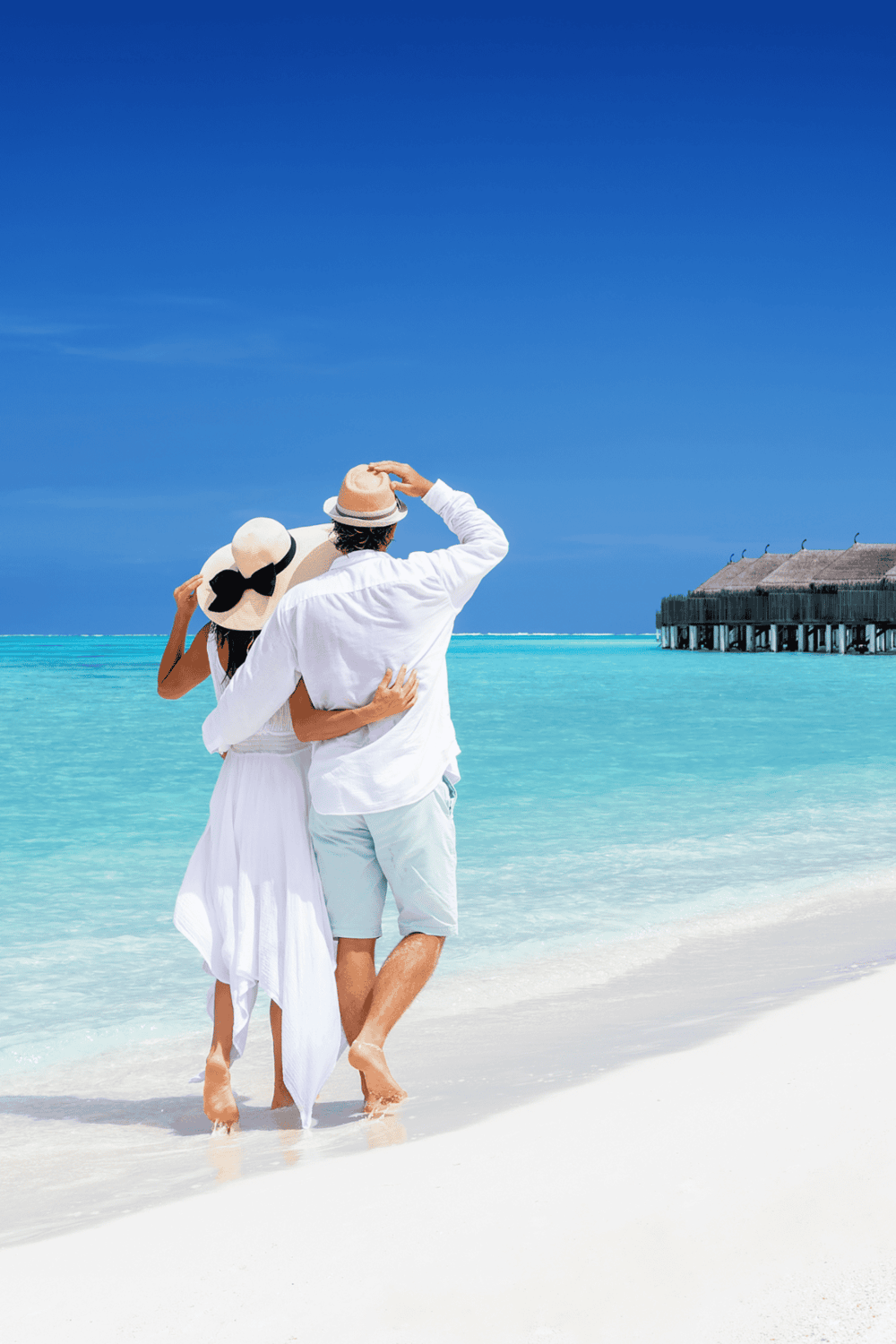 Relaxing couple walking on tropical beach with overwater bungalow in background.