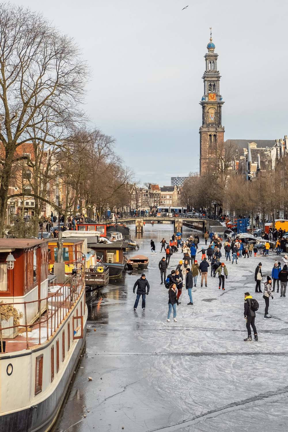 People skating on an icy canal in Amsterdam with the Montelbaanstoren tower in the background.