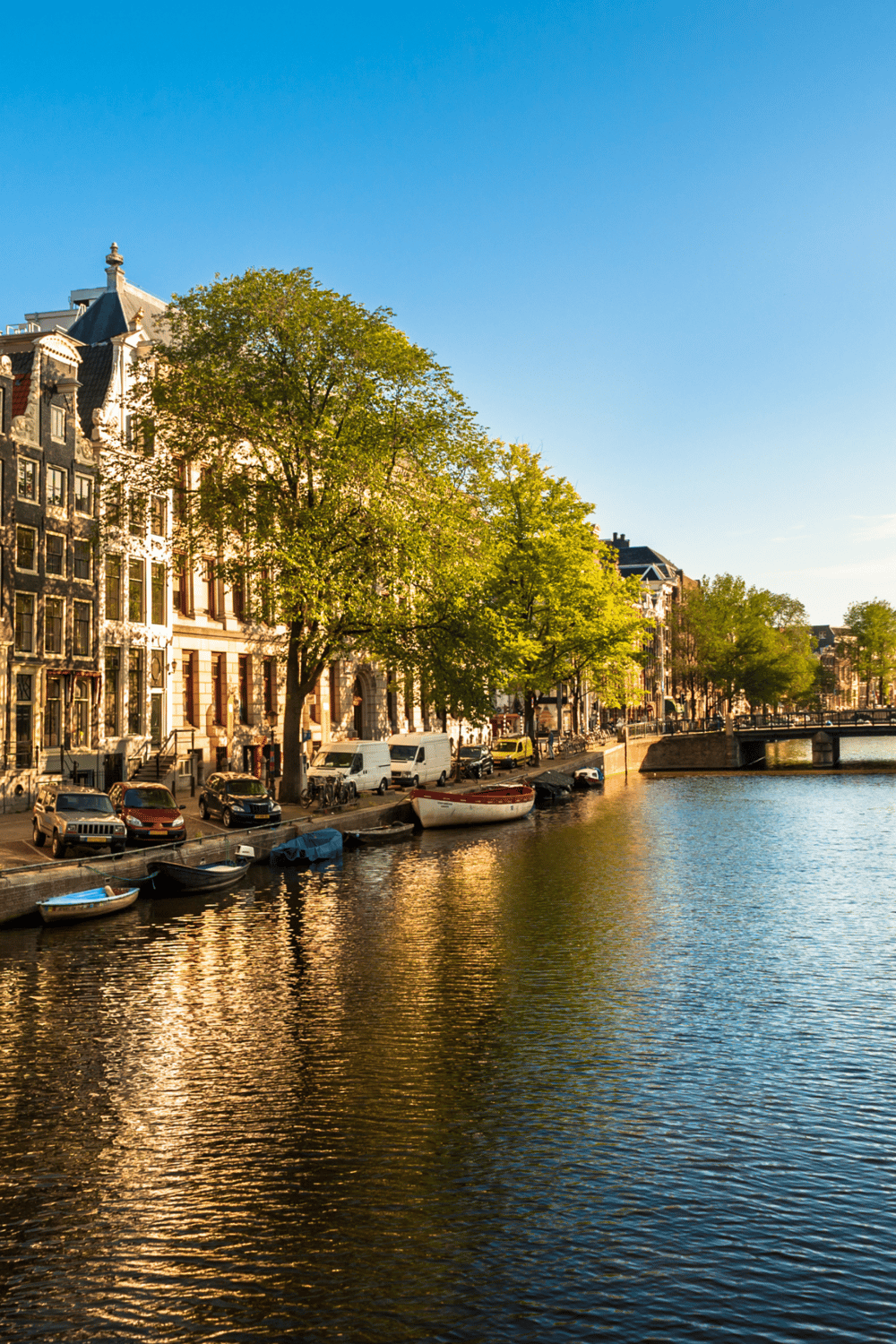 Boats docked along a scenic canal in Amsterdam with historic buildings and trees under a clear blue sky.