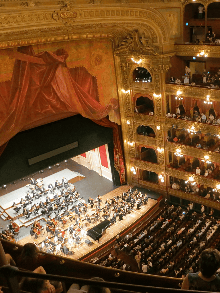 Grand opera house interior showcasing an orchestra and audience with ornate gold detailing and balconies.