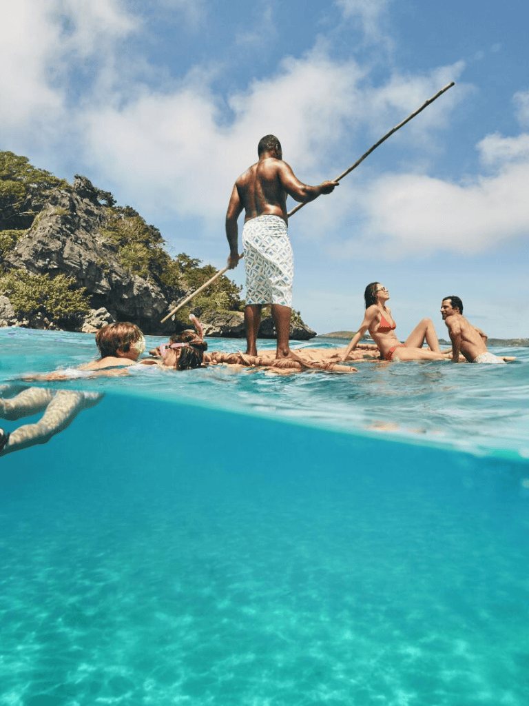 Traditional boat guide in tropical waters with tourists swimming and relaxing on a floating platform, scenic island background.