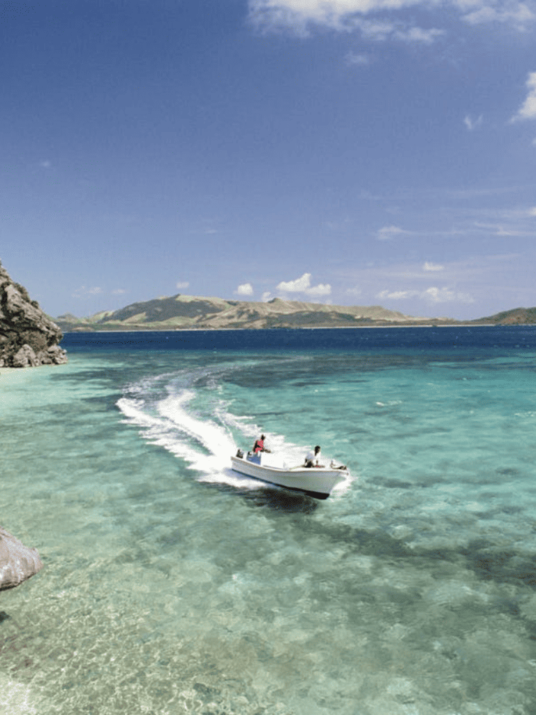 Seaside boat cruising over clear turquoise water near rocky coast and distant hills in sunny weather.