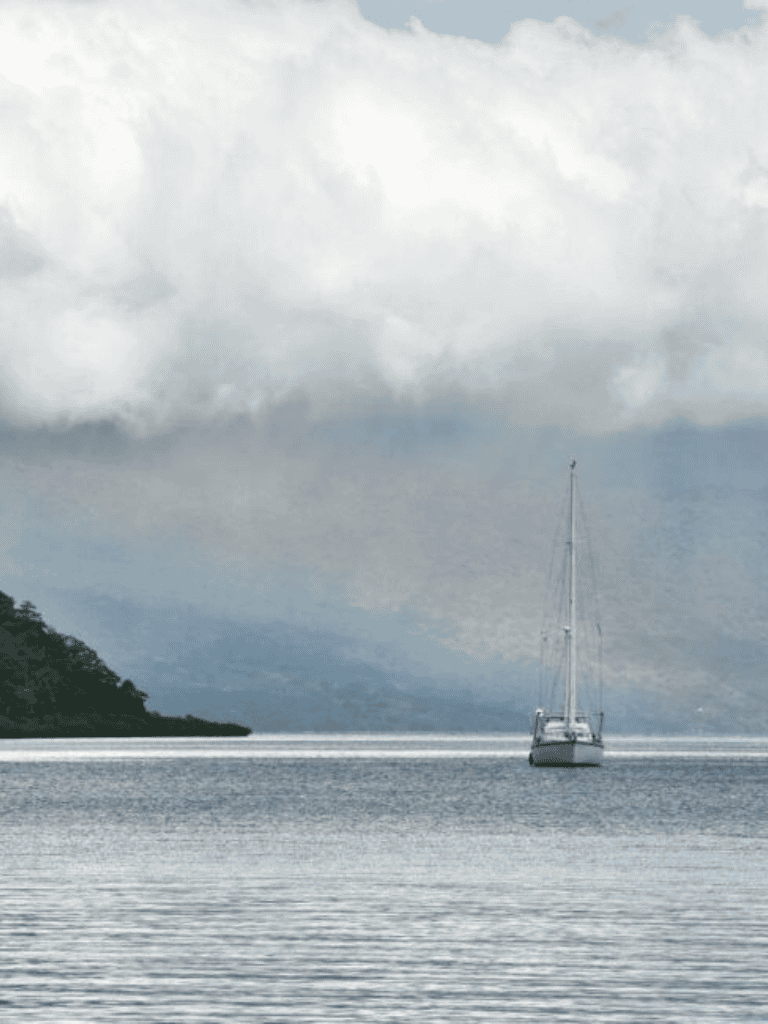 Serene sailboat on calm water under cloudy sky with lush island background.