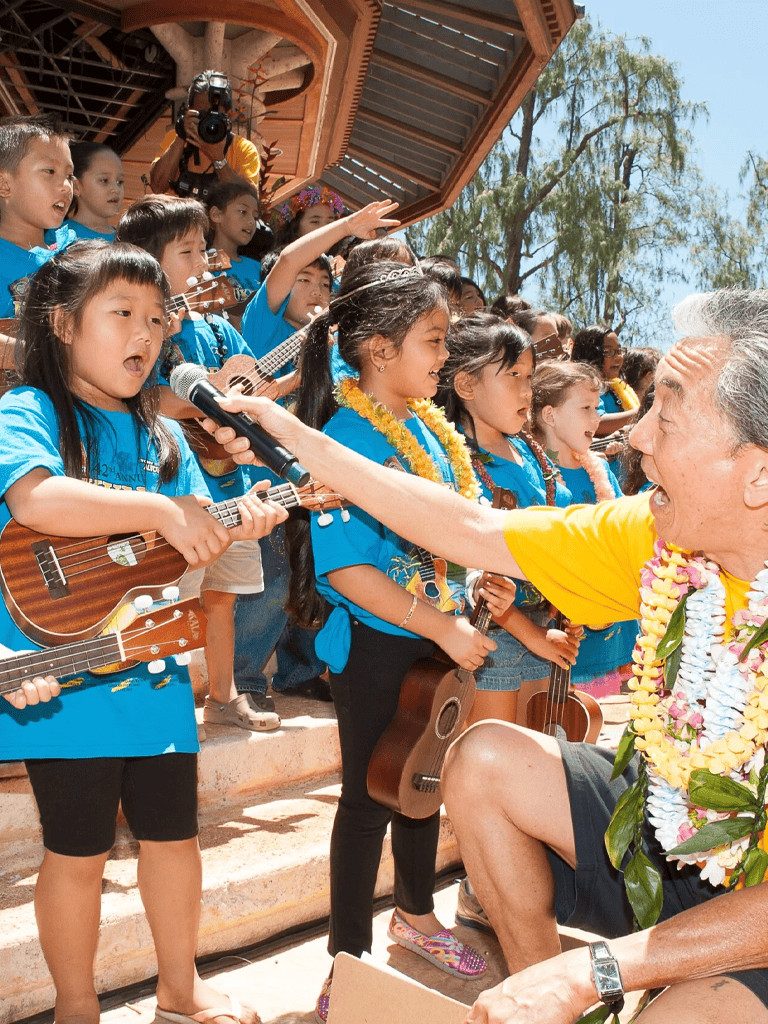 Joyful children performing with ukuleles at an outdoor cultural event, with an elderly man engaging with the kids.
