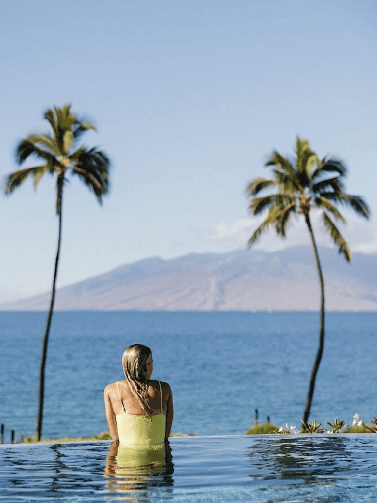 Relaxing woman in pool overlooking ocean with palm trees and mountain scenery in background.