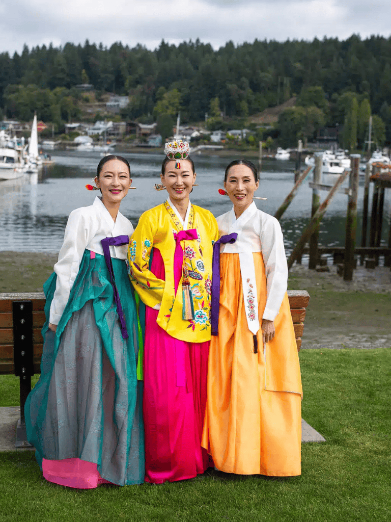 Traditional Korean women in colorful hanbok attire by the waterfront in a scenic outdoor setting.
