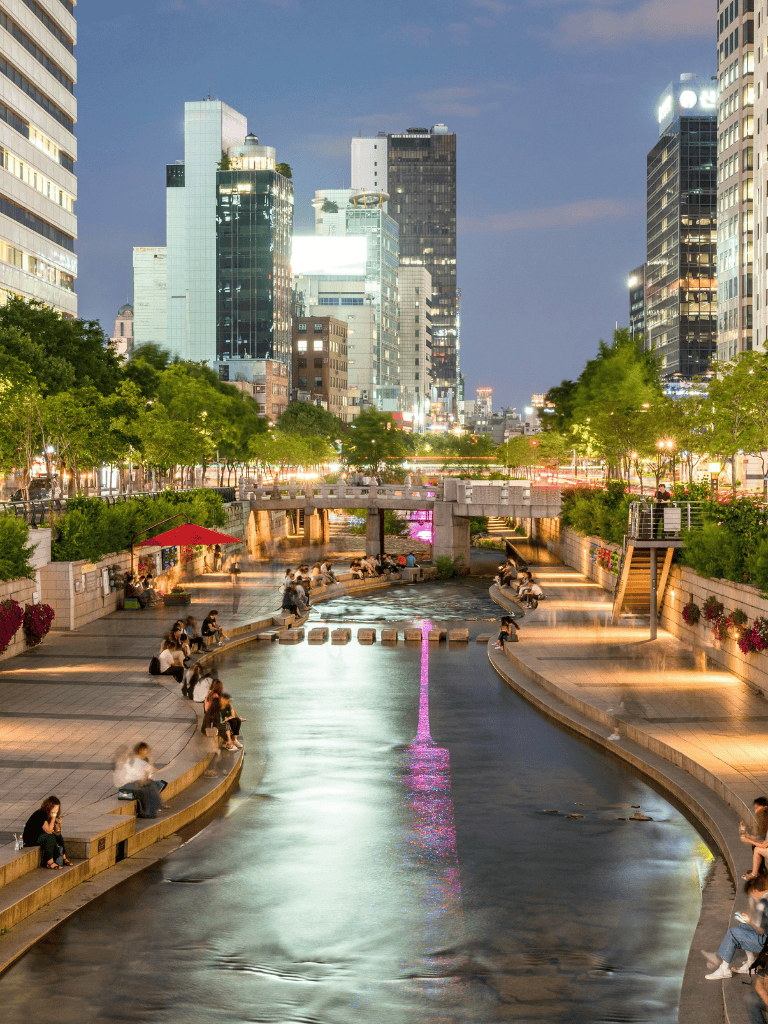 Plaza with cityscape and walking people, modern high-rise buildings, nighttime lighting, urban recreation area.