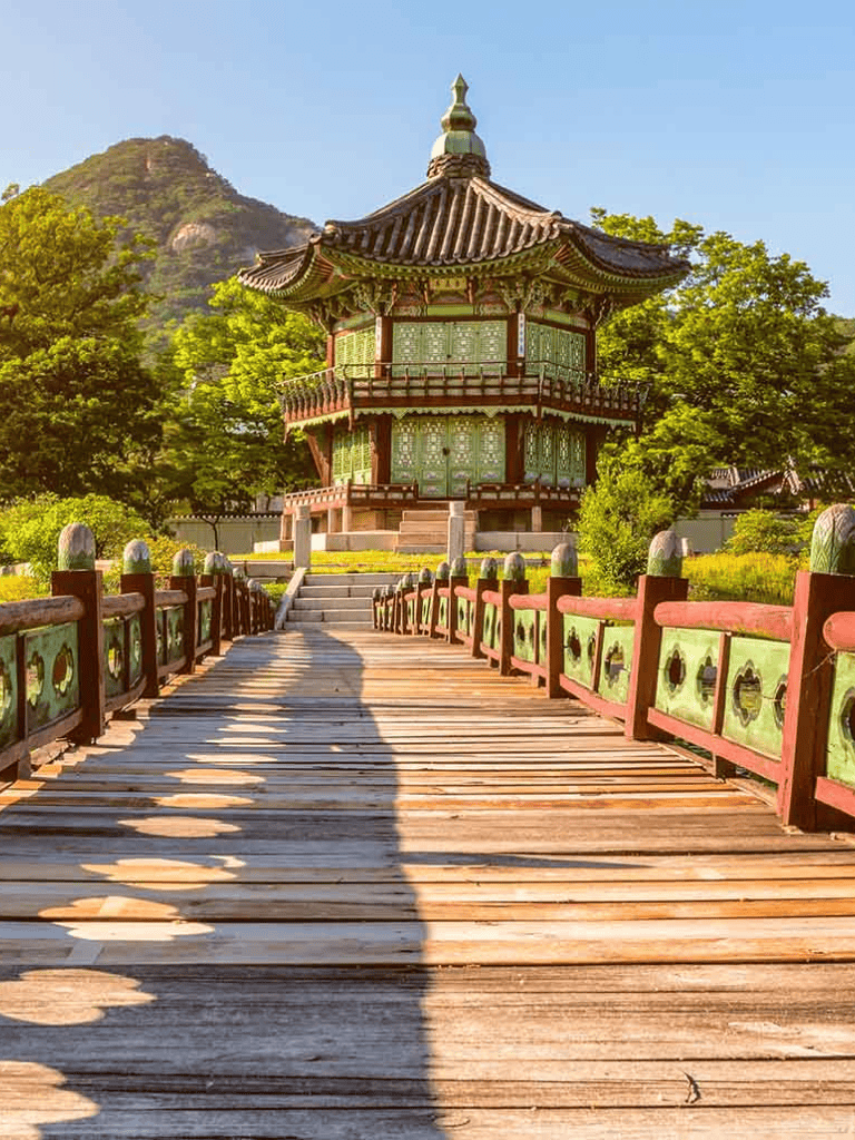 Colorful Korean Pavilion on a wooden bridge surrounded by lush greenery and mountain scenery.