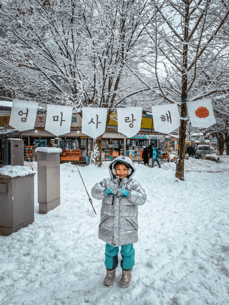 Snow-covered park with children enjoying winter, Korean sign banner, festive holiday atmosphere.